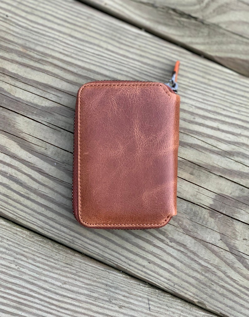 Puede incluir: Una cartera de cuero marr&oacute;n con cremallera y detalles de costura marr&oacute;n claro.