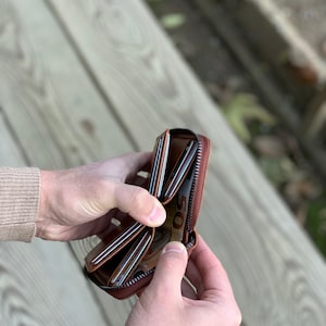 Puede incluir: Una cartera de cuero marr&oacute;n con cremallera, m&uacute;ltiples ranuras para tarjetas y un compartimento para billetes. La cartera est&aacute; abierta y muestra un billete de 50 euros en su interior.