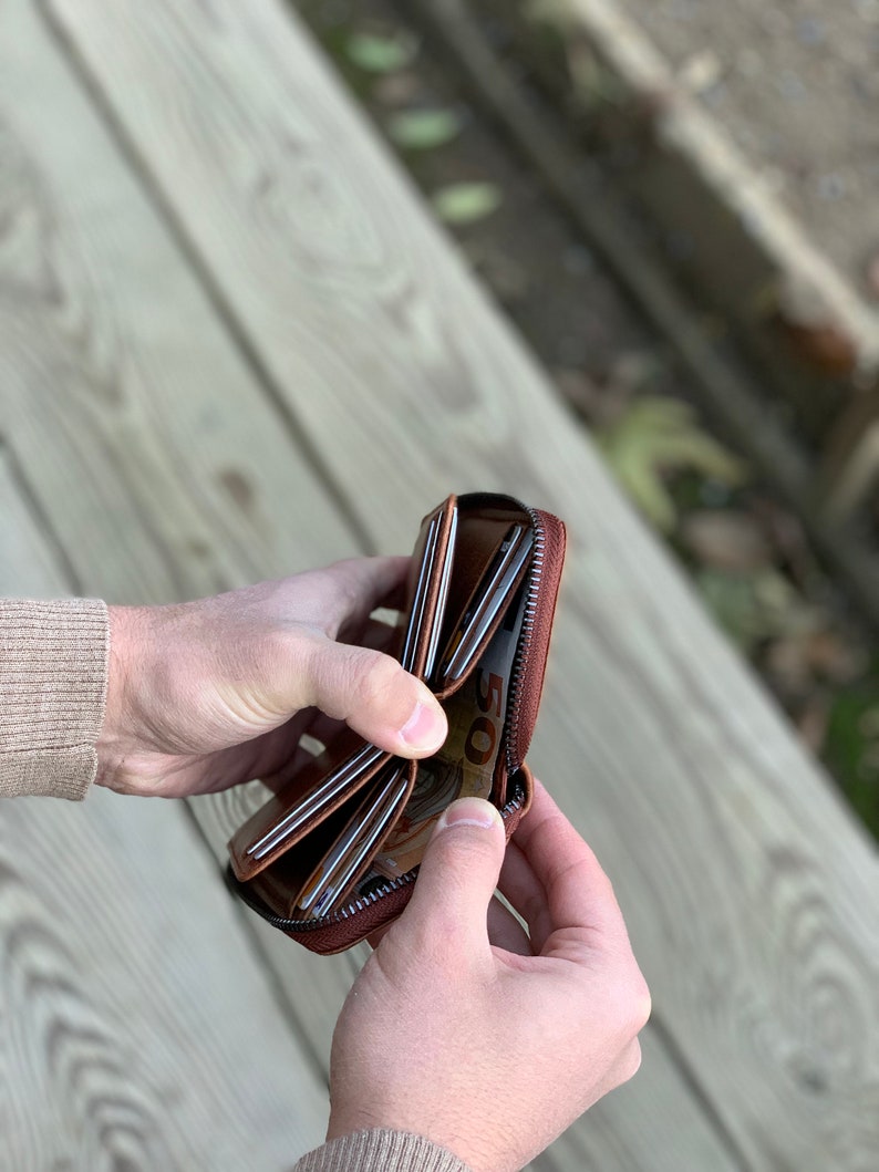 Puede incluir: Una cartera de cuero marr&oacute;n con cremallera, m&uacute;ltiples ranuras para tarjetas y un compartimento para billetes. La cartera est&aacute; abierta y muestra un billete de 50 euros en su interior.