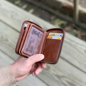 Puede incluir: Una cartera de cuero marr&oacute;n con cremallera, m&uacute;ltiples ranuras para tarjetas y una ventana transparente para el DNI. La cartera est&aacute; abierta y tiene varias tarjetas dentro.