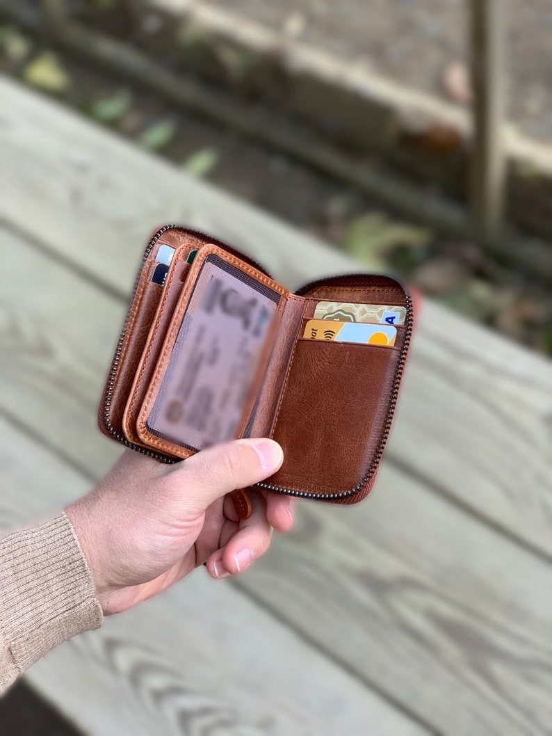 Puede incluir: Una cartera de cuero marr&oacute;n con cremallera, m&uacute;ltiples ranuras para tarjetas y una ventana transparente para el DNI. La cartera est&aacute; abierta y tiene varias tarjetas dentro.