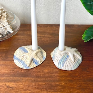 May include: Two ceramic candle holders with a seashell design. Each holder has a white candle. The holders are beige with blue accents and a starfish design. The holders are on a wooden surface.