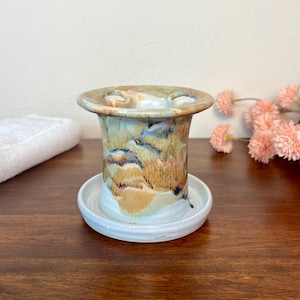 May include: A ceramic toothbrush holder featuring a flared top rim with two circular openings. The body displays a marbled glaze pattern in shades of tan, brown, blue, and cream, resting on a separate, solid white circular ceramic base tray.