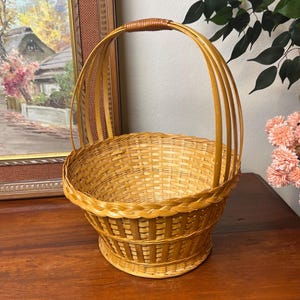 May include: A light brown wicker basket with a handle. The basket has a round base and a woven design. The handle is made of woven wicker and is arched. The basket is sitting on a wooden surface.