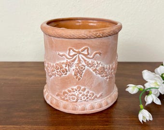 Puede incluir: Una maceta cilíndrica de terracota con un diseño en relieve de guirnaldas florales, lazos y un borde similar a una cuerda en la parte superior e inferior. El exterior tiene un acabado mate y terroso con una superficie interior lisa y esmaltada.