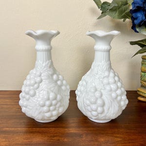Puede incluir: Dos jarrones blancos de vidrio opalino con dise&ntilde;o de racimo de uvas. Los jarrones tienen una parte superior con volantes y un cuerpo bulboso. Est&aacute;n sobre una superficie de madera oscura. Art&iacute;culos de decoraci&oacute;n.
