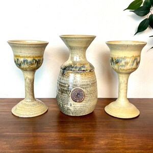 May include: Two ceramic goblets and a central vase featuring a speckled beige glaze with blue and brown drip patterns. The vase has a flared rim and a circular medallion detail on the body, while the goblets have tall stems and wide bases.