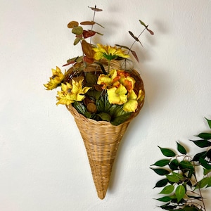 May include: A cone-shaped woven wicker wall basket filled with artificial yellow sunflowers, orange-tinted blossoms, and green eucalyptus-style foliage. The basket features a natural light brown finish and a tapered, pointed base design.