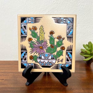 May include: Square ceramic tile featuring a Southwestern design. The tile has a cream-colored background with a central image of a cactus and flowers in a decorative pot. The border has geometric patterns in black, blue, and white. The tile is displayed on a black stand.