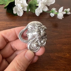 May include: A silver-coloured Art Nouveau brooch depicting a woman's profile with flowing hair and floral details. The brooch is held in a hand, with white flowers and green leaves in the background.