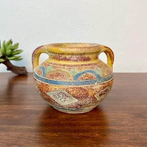 May include: A small, round ceramic vessel with two handles, featuring a textured, mottled finish in shades of yellow, red, and brown. The exterior is decorated with a repeating geometric pattern of blue arches and diamond shapes containing floral motifs.