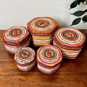 May include: A collection of five woven storage boxes with lids. The boxes are round and hexagonal, featuring a spiral design in shades of red, yellow, green, and brown. The boxes are arranged on a wooden surface.