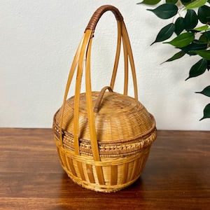 May include: A round, woven wicker basket with a lid and a handle. The basket is made of light brown wicker and has a darker brown handle. The handle is arched and made of woven material. The basket is sitting on a wooden surface.