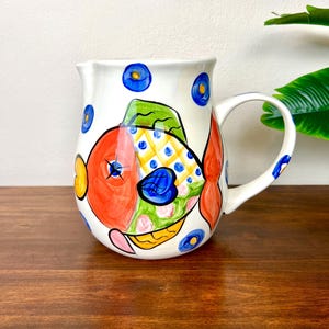 May include: A white ceramic pitcher featuring a hand-painted, colorful fish design. The fish has a large orange head, a yellow and blue checkered body, and green accents. The pitcher is decorated with small blue circles and has a curved handle.