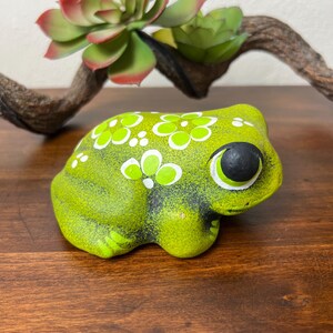 May include: A ceramic frog figurine in shades of green, decorated with white floral patterns. The frog has large, black-pupiled eyes and is positioned on a wooden surface. A decorative succulent and branch are in the background.