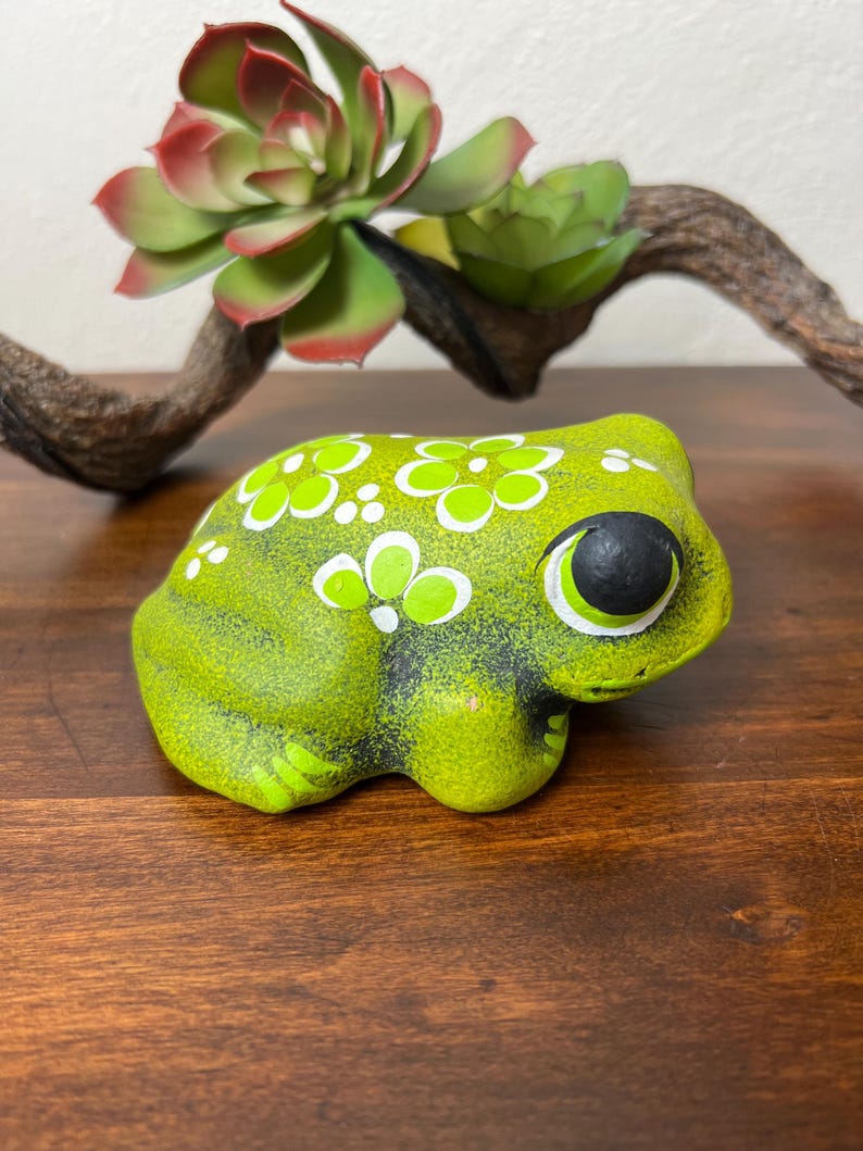 May include: A ceramic frog figurine in shades of green, decorated with white floral patterns. The frog has large, black-pupiled eyes and is positioned on a wooden surface. A decorative succulent and branch are in the background.