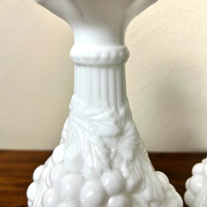 Puede incluir: Candelabros blancos de estilo vintage con un dise&ntilde;o de uvas y hojas. Los candelabros tienen una parte superior acampanada y una superficie texturizada detallada. La base est&aacute; decorada con un racimo de uvas.