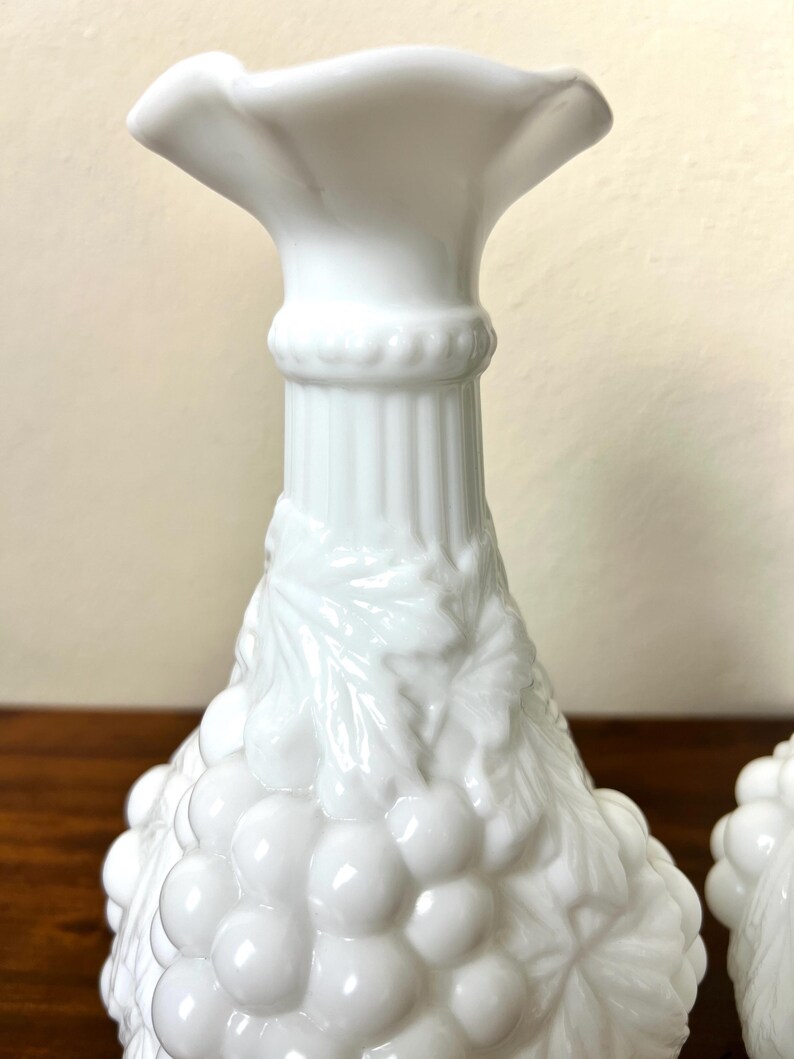 Puede incluir: Candelabros blancos de estilo vintage con un dise&ntilde;o de uvas y hojas. Los candelabros tienen una parte superior acampanada y una superficie texturizada detallada. La base est&aacute; decorada con un racimo de uvas.