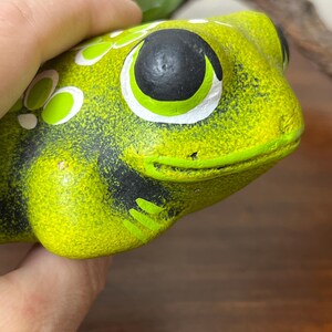 May include: A ceramic frog figurine in shades of green and black. The frog has large, round eyes with black pupils and white and green accents. The frog's body is a textured, lime green color with black markings.