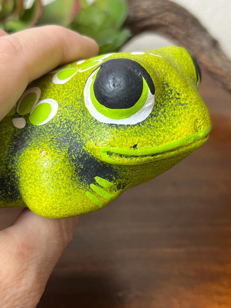 May include: A ceramic frog figurine in shades of green and black. The frog has large, round eyes with black pupils and white and green accents. The frog's body is a textured, lime green color with black markings.
