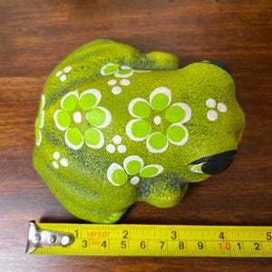 May include: A ceramic frog figurine in shades of green, adorned with white and light green floral patterns. The frog has black eyes and a black mouth. The figurine is approximately 4 inches long.