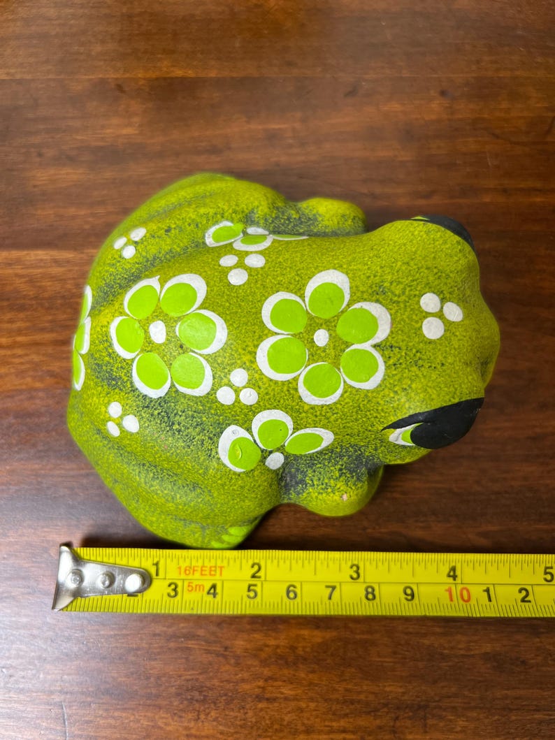May include: A ceramic frog figurine in shades of green, adorned with white and light green floral patterns. The frog has black eyes and a black mouth. The figurine is approximately 4 inches long.