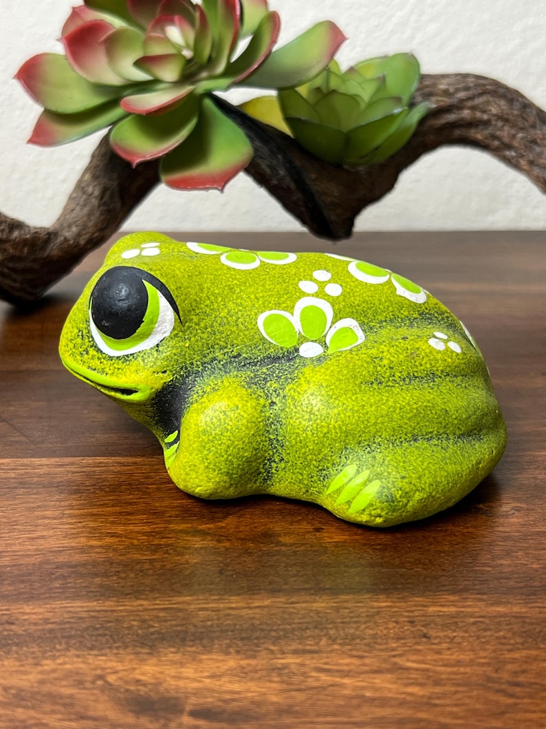 May include: A ceramic frog figurine in shades of green, black, and white. The frog has large eyes and is decorated with white floral patterns. The figurine is on a wooden surface, with a succulent plant in the background.