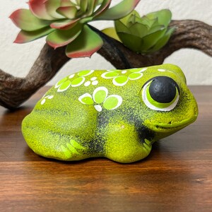 May include: A ceramic frog figurine in shades of green, with white and lime green floral accents. The frog has large, expressive eyes and sits on a wooden surface. Artificial succulents and a branch are in the background.