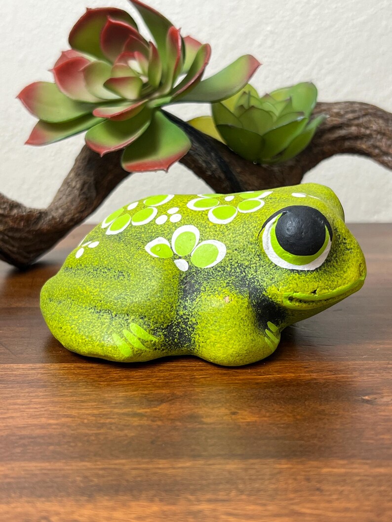 May include: A ceramic frog figurine in shades of green, with white and lime green floral accents. The frog has large, expressive eyes and sits on a wooden surface. Artificial succulents and a branch are in the background.