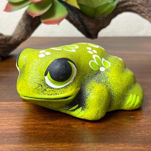 May include: A lime green ceramic frog figurine with black accents around the eyes and mouth. White floral designs are painted on the frog's back. The frog is sitting on a wooden surface.