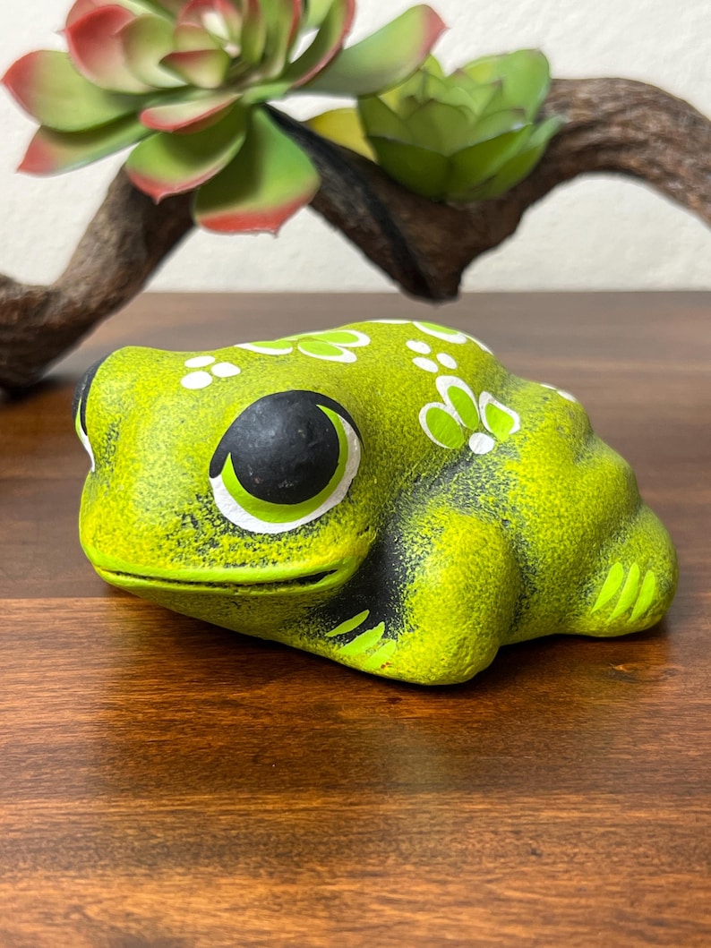May include: A lime green ceramic frog figurine with black accents around the eyes and mouth. White floral designs are painted on the frog's back. The frog is sitting on a wooden surface.