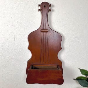 May include: A wooden wall-mounted violin-shaped shelf with a dark brown finish. The shelf features carved details mimicking a violin's strings and tuning pegs. A small, open box is at the bottom. The shelf is hanging on a white wall.