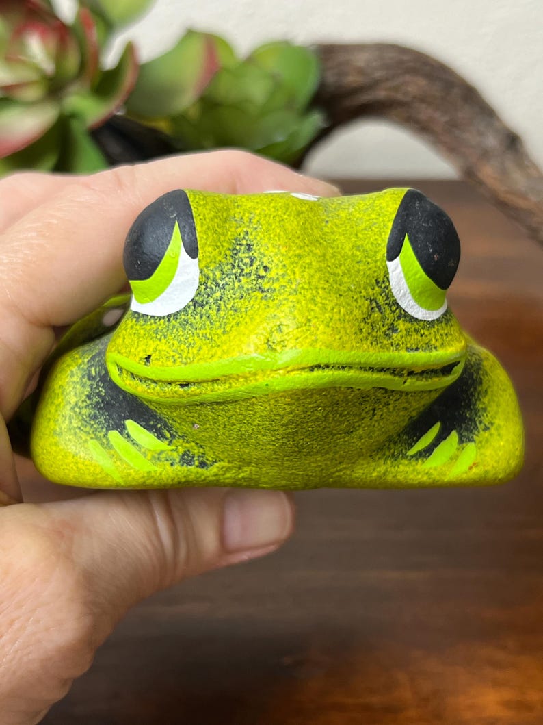 May include: A ceramic frog figurine in shades of green, black, and white. The frog has large, expressive eyes and a textured surface. The figurine is held in a hand, with a blurred background of greenery and wood.