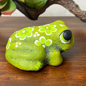 May include: A ceramic frog figurine in shades of green, decorated with white floral patterns. The frog has large, black eyes with white accents. The figurine is placed on a wooden surface.