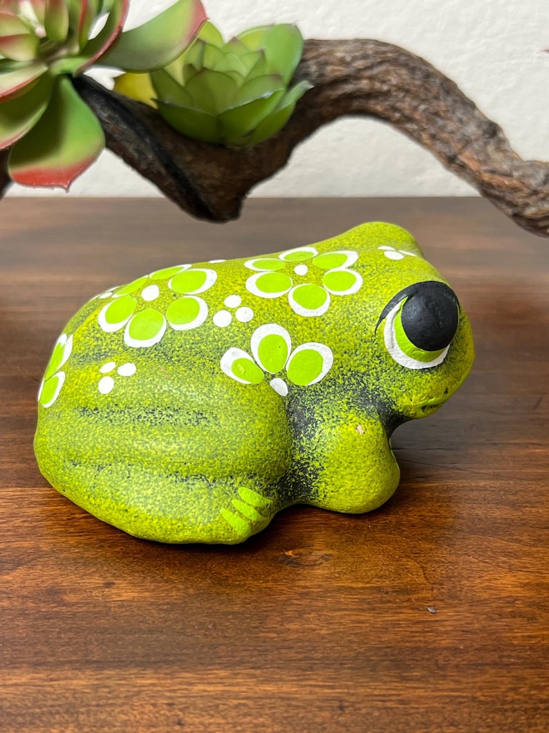 May include: A ceramic frog figurine in shades of green, decorated with white floral patterns. The frog has large, black eyes with white accents. The figurine is placed on a wooden surface.