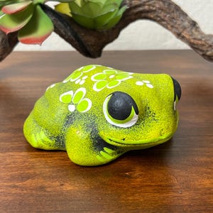 May include: A ceramic frog figurine in shades of green and yellow, with black eyes and white floral accents. The frog is sitting on a wooden surface, with a succulent and a piece of driftwood in the background. The figurine is a decorative item.