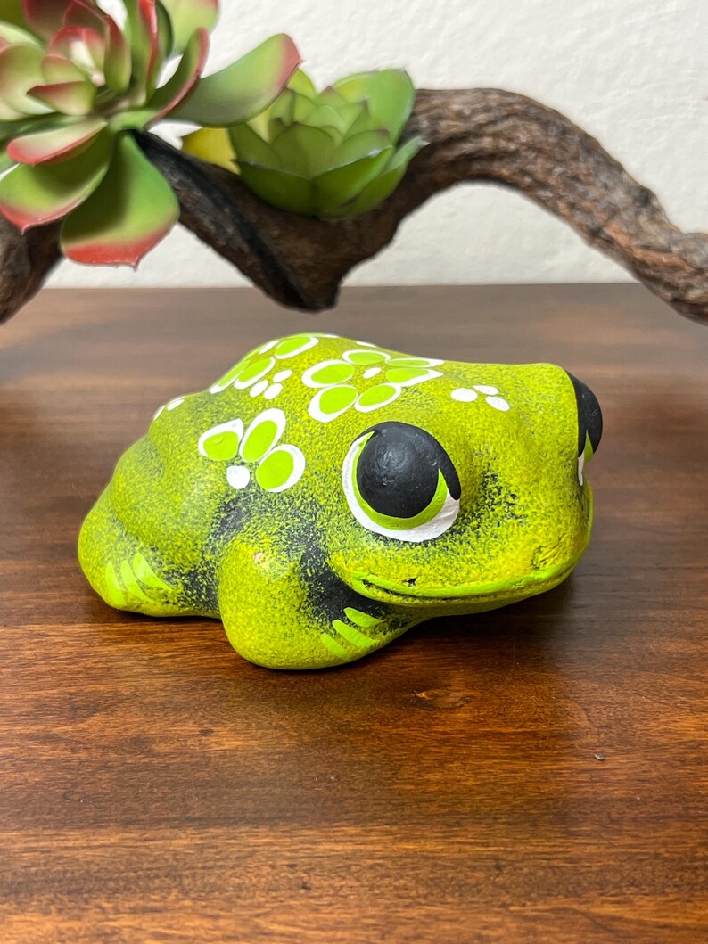May include: A ceramic frog figurine in shades of green and yellow, with black eyes and white floral accents. The frog is sitting on a wooden surface, with a succulent and a piece of driftwood in the background. The figurine is a decorative item.