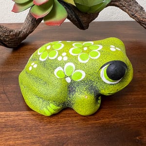 May include: A ceramic frog figurine in shades of green, with white and green floral accents. The frog has a black eye and is sitting on a wooden surface. A faux succulent and branch are in the background.