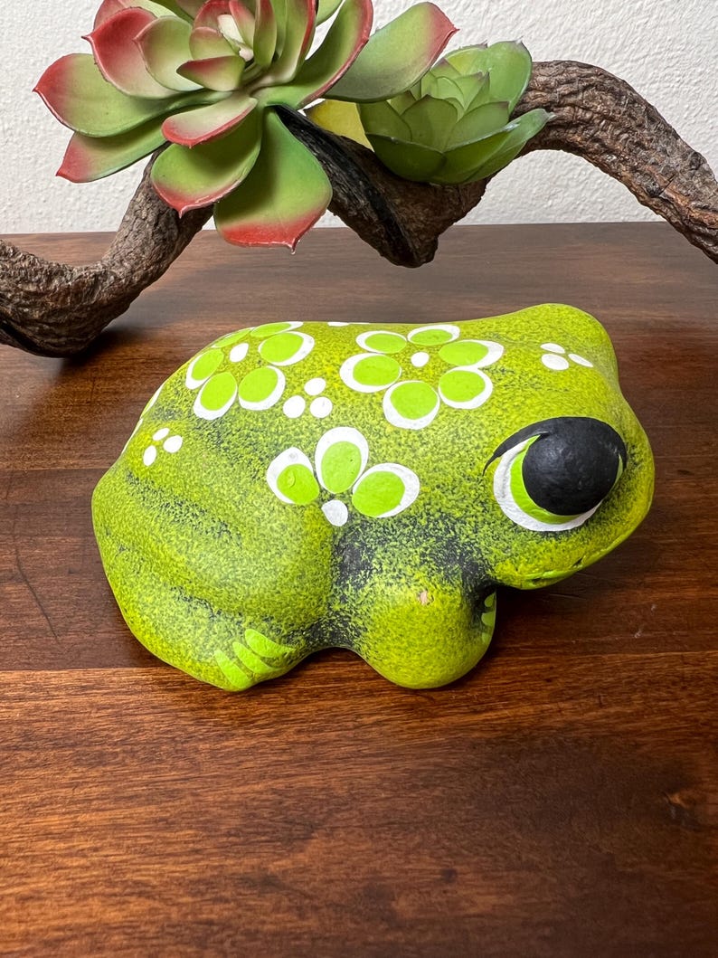 May include: A ceramic frog figurine in shades of green, with white and green floral accents. The frog has a black eye and is sitting on a wooden surface. A faux succulent and branch are in the background.