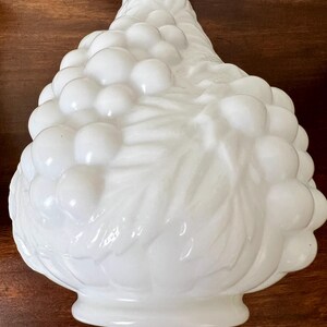 Puede incluir: Un jarr&oacute;n decorativo de vidrio blanco con un dise&ntilde;o detallado de racimo de uvas. El jarr&oacute;n tiene una forma bulbosa con una base acampanada y un cuello delgado. La superficie es lisa y brillante, reflejando la luz.