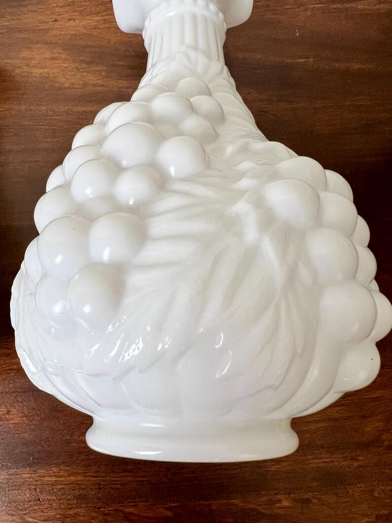 Puede incluir: Un jarr&oacute;n decorativo de vidrio blanco con un dise&ntilde;o detallado de racimo de uvas. El jarr&oacute;n tiene una forma bulbosa con una base acampanada y un cuello delgado. La superficie es lisa y brillante, reflejando la luz.