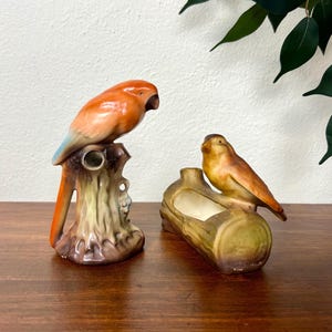 Op de afbeelding: Twee keramische vogelbeeldjes. Eén is een papegaai op een boomstronk, met oranje, blauwe en bruine tinten. De andere is een kleine vogel op een boomstamvormige plantenbak, in bruine en gele tinten. Beide zijn decoratieve items.