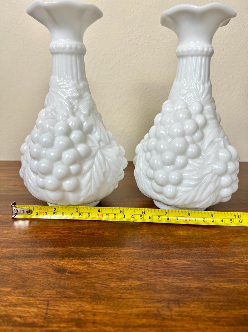 Puede incluir: Dos jarrones blancos de vidrio opalino. Cada jarr&oacute;n presenta un cuerpo bulboso con un dise&ntilde;o de racimo de uvas y una parte superior con volantes. Los jarrones miden aproximadamente 25 cm de alto, con una cinta m&eacute;trica en primer plano.