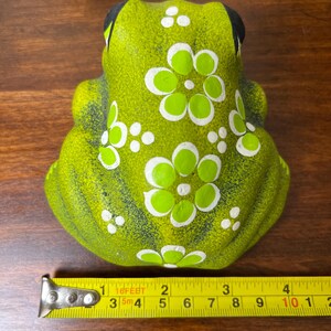 May include: A green ceramic frog figurine with white and green floral designs. The frog has black eyes and is approximately 4 inches long. The figurine is on a wooden surface.