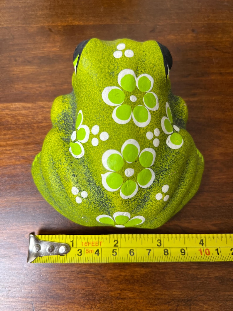 May include: A green ceramic frog figurine with white and green floral designs. The frog has black eyes and is approximately 4 inches long. The figurine is on a wooden surface.