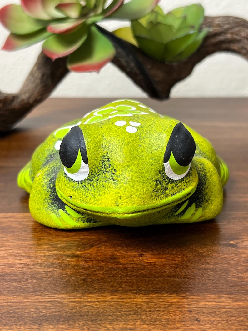 May include: A ceramic frog figurine in shades of green, with black and white accents. The frog has a smiling expression and is decorated with white dots. The figurine is on a wooden surface, with a succulent and branch in the background.