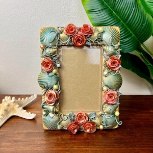 Könnte beinhalten: Rechteckiger Fotorahmen mit Muschel- und Rosenmotiv. Der Rahmen ist mit türkisfarbenen Muscheln, korallenfarbenen Rosen und Kunstperlen verziert. Der Rahmen hat einen goldfarbenen Rand und eine braune Rückseite.