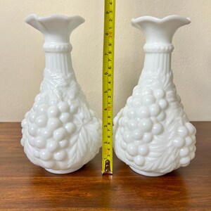 Puede incluir: Dos jarrones de vidrio blanco lechoso. Cada jarr&oacute;n tiene una parte superior acampanada, un cuello acanalado y un cuerpo decorado con un dise&ntilde;o de racimo de uvas. Los jarrones miden aproximadamente 20 cm de altura.