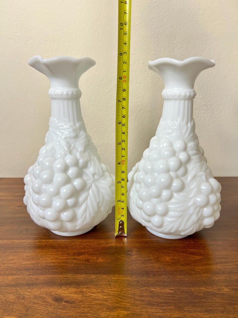 Puede incluir: Dos jarrones de vidrio blanco lechoso. Cada jarr&oacute;n tiene una parte superior acampanada, un cuello acanalado y un cuerpo decorado con un dise&ntilde;o de racimo de uvas. Los jarrones miden aproximadamente 20 cm de altura.