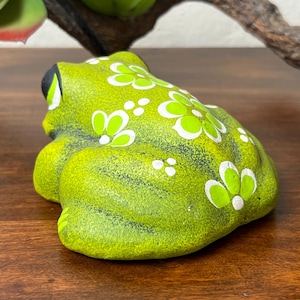 May include: A ceramic frog figurine in a vibrant green hue, adorned with white and lime green floral patterns. The frog has black eyes and is positioned on a wooden surface. A decorative item for home decor.